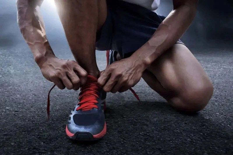 A person kneels on a dark surface, tying the laces of a sporty shoe with red accents.