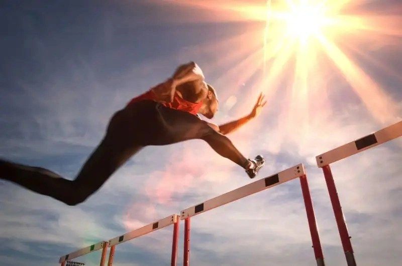 An athlete hurdles over a barrier under a bright sun, showcasing dynamic movement against a blue sky.