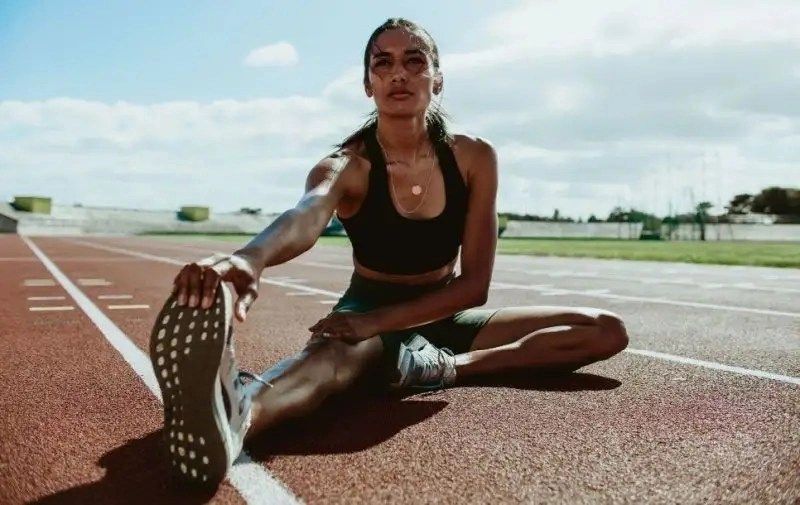 A woman stretches on a running track, wearing athletic gear, with a focus on her leg and foot. Bright sky in the background.