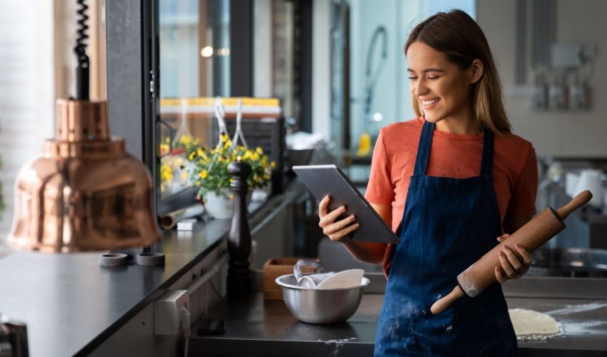 Cheerful female food blogger professional baker looking at digital tablet in commercial kitchen.