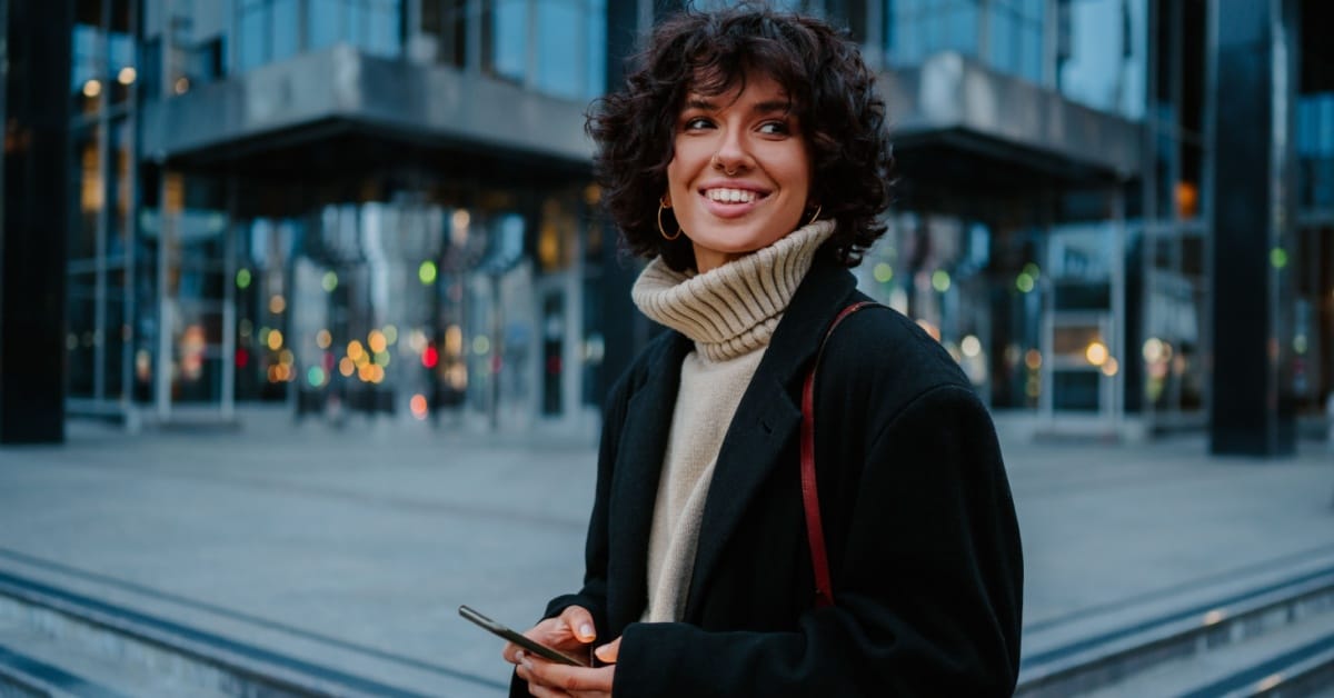 A smiling young woman holding her smart phone while waiting in front of a building.